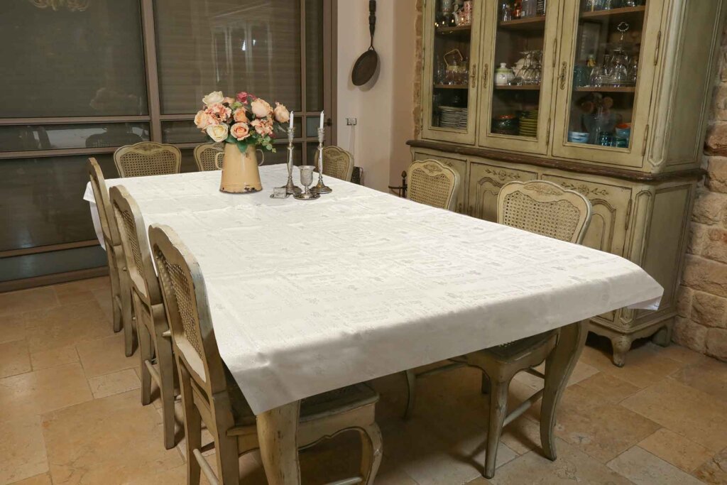 מפת שולחן מלבנית מעוטרת בדוגמה עדינה פרוסה על שולחן אוכל חגיגי מוקף כיסאות בחדר אוכל בסגנון כפרי